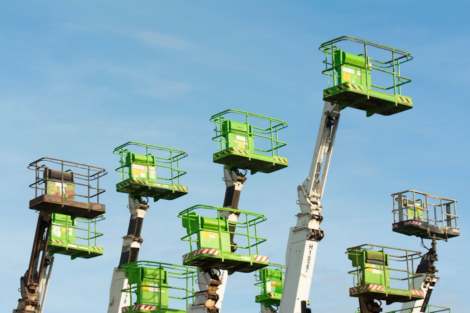 A group of green cherry pickers extending against a clear blue sky in an industrial setting.